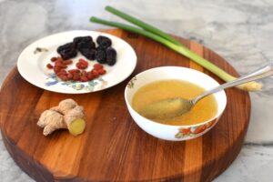 A bowl of herbal chicken soup, a healing postpartum soup recipe with red dates, ginger, goji berries, and scallion on the side.