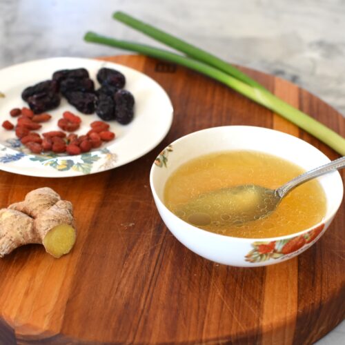 A bowl of herbal chicken soup, a healing postpartum soup recipe with red dates, ginger, goji berries, and scallion on the side.